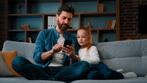 Happy young girl and father look at smartphone on sofa