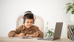 Smiling caucasian little kid in headphones sits at table, makes notes, watches lesson on computer in room interior. Study, education at school, kindergarten and home remotely, childhood and video call