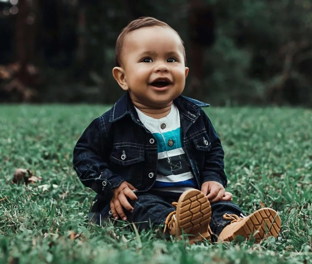 one year old boy in denim jeans and jacket, sat in a field about with mouth movement indicating he's about to speak.