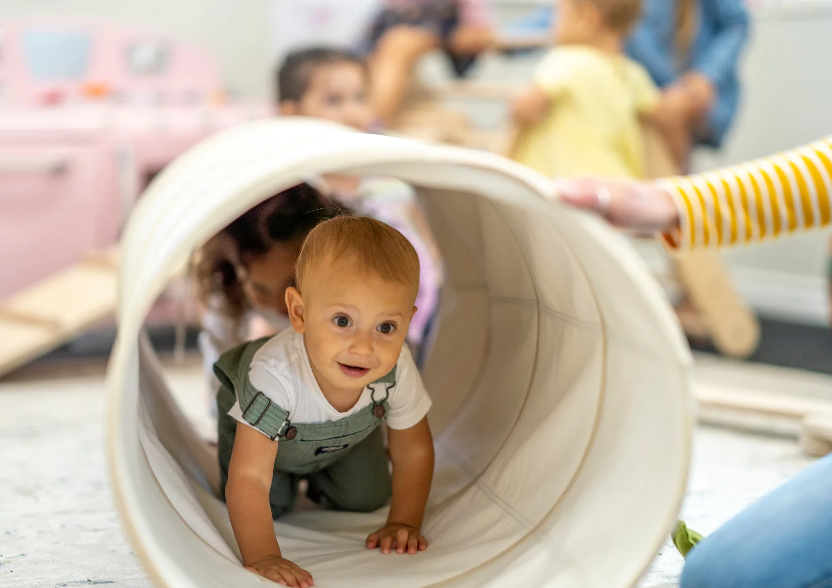 Baby in daycare with caregiver, smiling crawling through tunnel